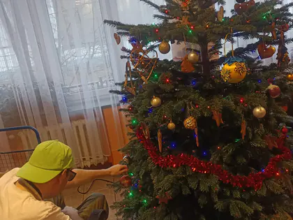 Ubrana choinka z lampkami i bombkami, przy niej mężczyzna w zielonej czapce