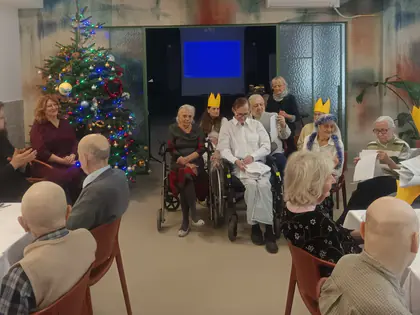 Mieszkańcy siedzą w jadalni przy choince, część w papierowych koronach, z boku ksiądz i dwie osoby prowadzące spotkanie