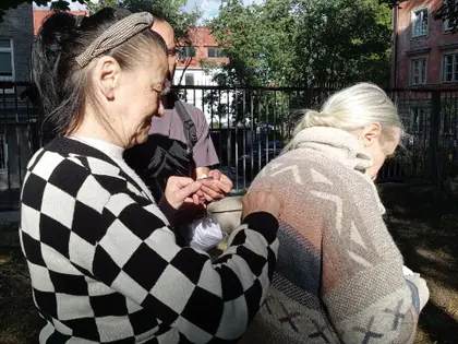 Starsza kobieta w beżowym swetrze odwrócona tyłem, za nią kobieta w swetrze w szachownicę, w tle zieleń ogrodu