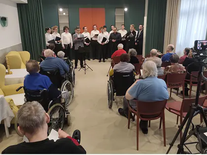 Chór w białych koszulach śpiewa na sali, przed nim siedzą słuchacze, z boku kamera na statywie
