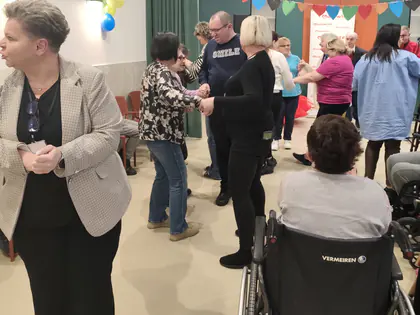 Kobieta w jasnej marynarce stoi na sali, za nią tańczą pary i siedzi osoba na wózku