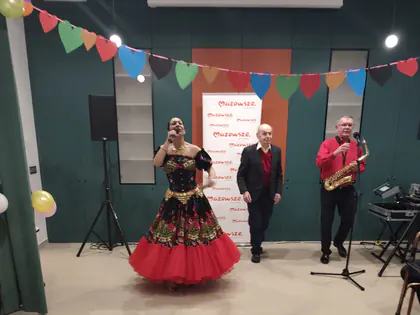 Wokalistka w czarno-czerwonej sukni i saksofonista przy mikrofonach, obok stoi starszy mężczyzna w ciemnej marynarce