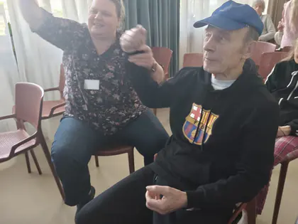 Starszy mężczyzna w niebieskiej czapce trzyma za rękę siedzącą obok kobietę, która tanecznie unosi rękę