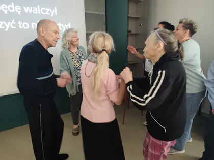 Taneczny krąg starszych osób trzymających się za ręce, w tle biały ekran z tekstem piosenki