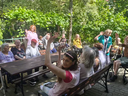 Starsze osoby siedzą radośnie przy stole, z tyłu kobieta w różowej koszuli wesoło macha rękami, w tle ogród