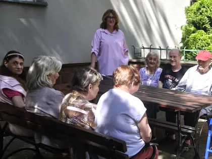 Radosna grupa osób przy drewnianym stole patrzy na stojącą kobietę w różowej koszuli