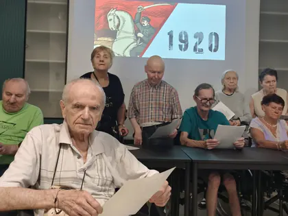 Starszy mężczyzna czyta wiersz, w tle inne osoby przy stole z kartkami wierszy