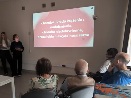 Seniorzy patrzą na slajd o chorobach układu krążenia wyświetlany na ścianie, obok stoją dwie studentki prowadzące wykład
