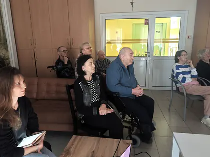Seniorzy w świetlicy słuchają wykładu, z przodu siedzi studentka z tabletem
