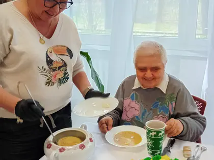 Kobieta nalewa zupę starszej pani siedzącej przy stole
