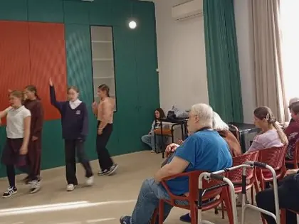 Grupa taneczna młodych osób, jedna podnosi rękę, druga śpiewa, z przodu siedzą mężczyzna i starsza kobieta i przyglądają się scenie