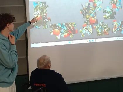 Młody mężczyzna wskazuje fragment układanki wyświetlanej na dużym ekranie, obok siedzi starszy mężczyzna na wózku