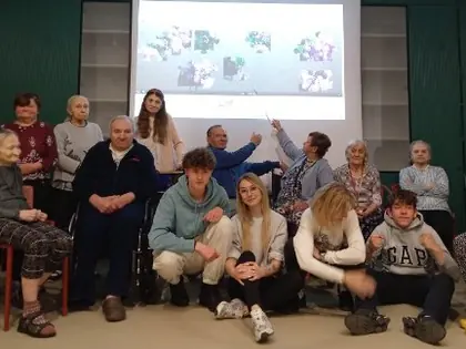 Grupa osób siedzi obok siebie, z tyłu dwie osoby wskazują na ekran