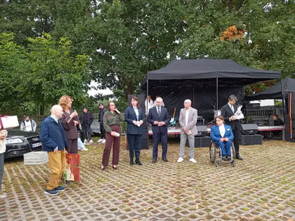 Kobieta przemawia do osób stojących w szeregu, obok mężczyzna w średnim wieku trzyma obraz, w tle sprzęt muzyczny