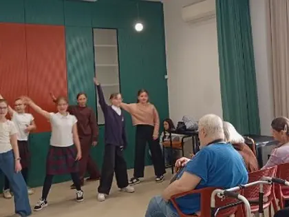 Grupa młodych osób tańczy z podniesionymi rękami, przed nimi na czerwonych krzesłach siedzą starszy mężczyzna i kobieta i przyglądają się występowi