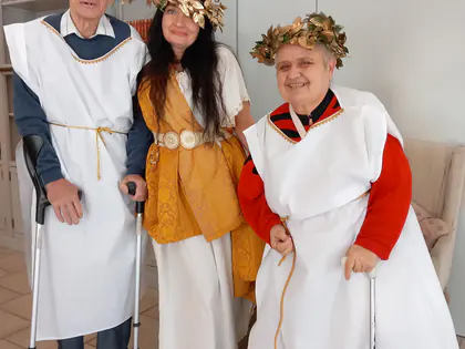 Troje uśmiechniętych osób w białych tunikach i złotych wieńcach laurowych, dwie z nich opierają się na kulach