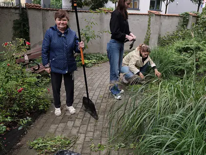 Dwie wolontariuszki pielą rośliny, z przodu pani w niebieskiej kurtce trzyma szpadel, w tle ogród