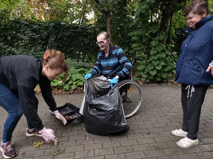 W ogrodzie pochylona wolontariuszka sprząta liście, obok mężczyzna na wózku trzyma czarny worek, z boku przygląda się kobieta w niebieskiej kurtce