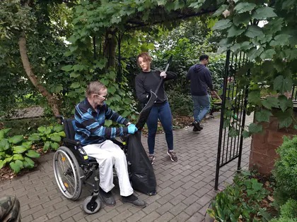Mężczyzna na wózku trzyma czarny worek ogrodowy, do którego wolontariuszka wrzuca szuflą liście, w tle ogród i drugi pracujący mężczyzna