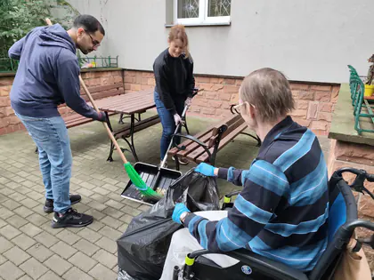 Osoby sprzątają ogród, mężczyzna na wózku trzyma worek na liście, wolontariusze zgarniają liście na szuflę, w tle ławki ogrodowe