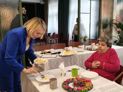 Kobieta podaje posiłek starszej pani, w tle długi stół z potrawami i wielkanocnym wystrojem